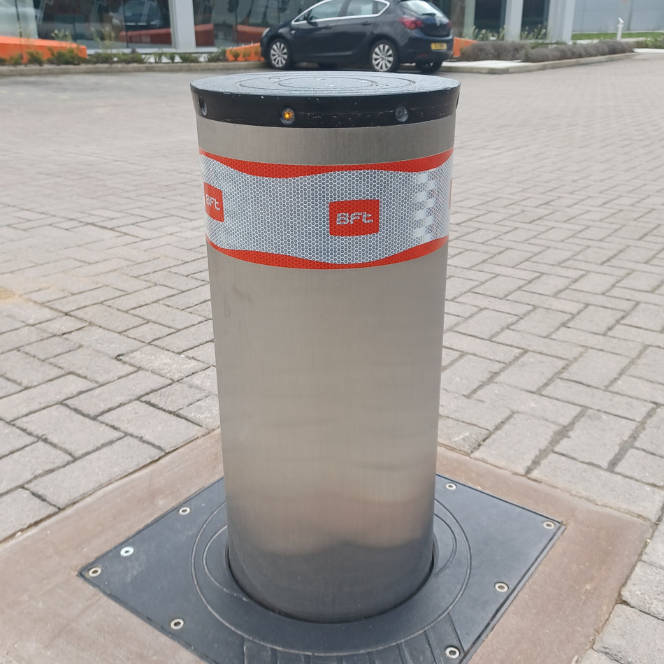 A stainless steel cylindrical automatic rising bollard with a reflective band featuring the red BFT logo, installed on a paved outdoor driveway.
