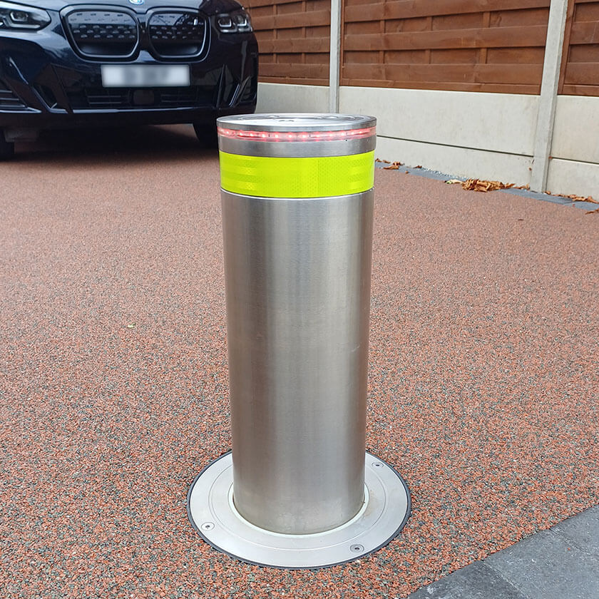 Stainless steel automatic rising bollard  with a Yellow reflective band on a driveway with a car in the background.