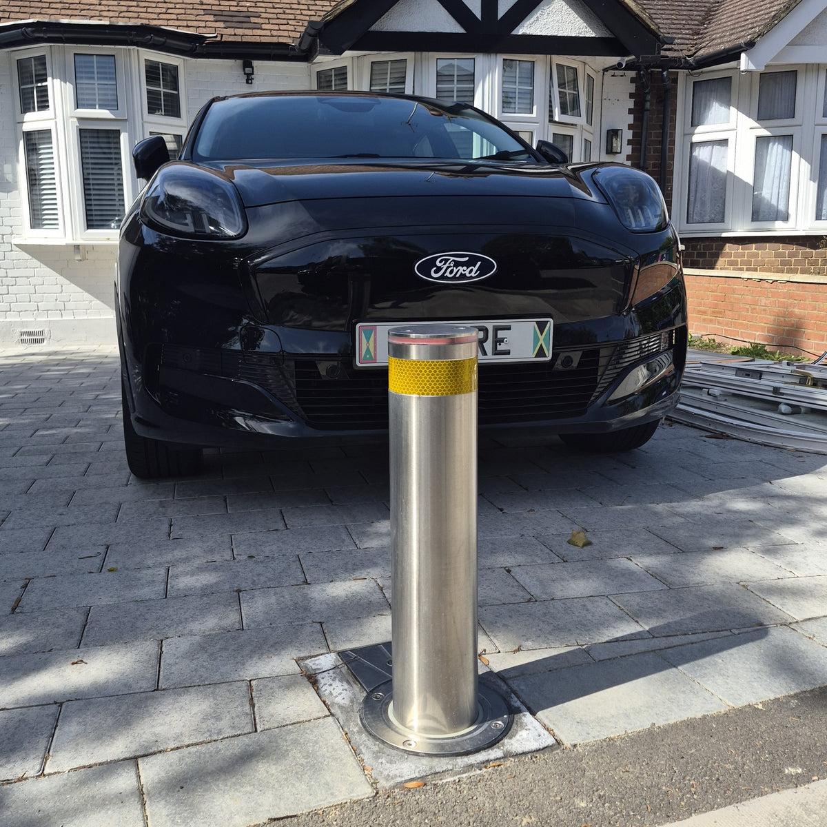 A silver metal automatic bollard with a yellow reflective band near the top, installed on a paved driveway in front of a black Ford car.