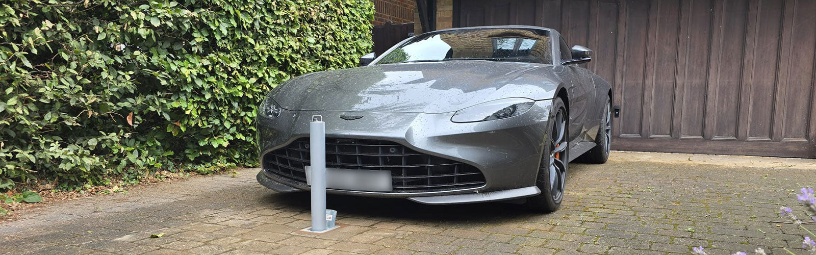 High security telescopic bollard installed in front of a Grey sports car parked on a driveway next to a wooden fence and greenery.