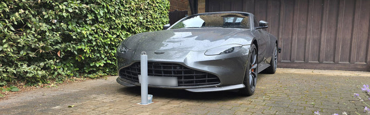 High security telescopic bollard installed in front of a Grey sports car parked on a driveway next to a wooden fence and greenery.