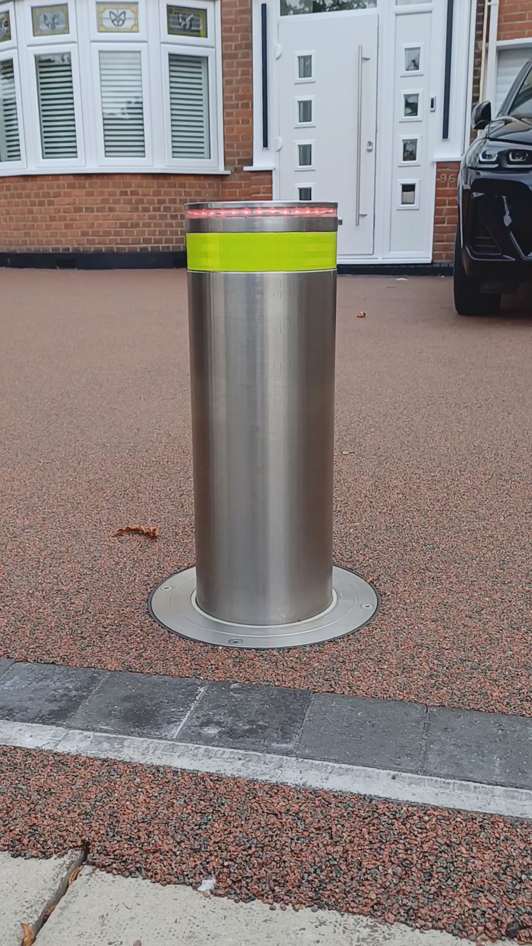 Stainless steel automatic rising bollard  with a Yellow reflective band on a driveway with a car in the background.