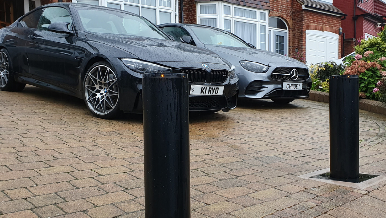 Load video: A black BMW M4 coupe parked on a paved residential driveway, with a silver Mercedes-Benz car behind it and black automatic bollards in the foreground.
